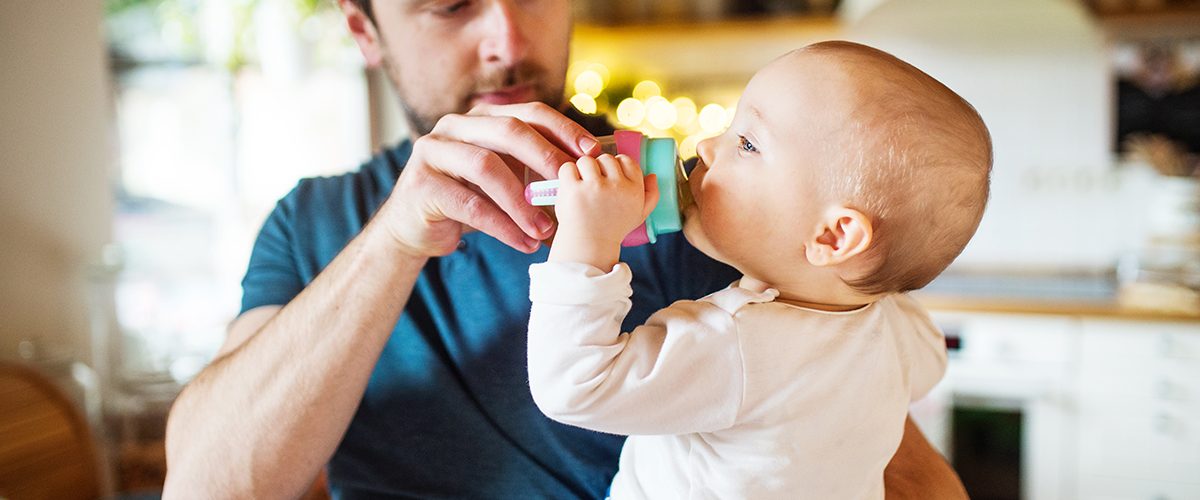 A father with a baby daughter at home. A cute girl drinking water from the bottle. Paternity leave.